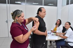 Homem de camisa preta sendo vacinado por enfermeira vestida de blusa na cor vinho e duas enfermeiras sentadas em uma mesa a sorrir